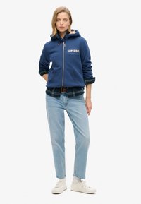 Sudadera con cremallera color azul marino, con bolsillos frontales, logo bordado, superpuesta sobre una camisa de cuadros, combinada con jeans de color azul claro y zapatillas blancas.