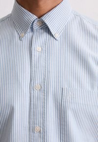 Camisa de botones a rayas azules y blancas con cuello clásico, que presenta un bolsillo en el pecho izquierdo y botones lisos. Tela de algodón texturizada.