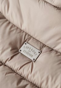 Szczegół kurtki pikowanej w miękkim beżu z subtelną teksturą, z metalową etykietą marki w kolorze srebrnym, na której widnieje napis "SUPERDRY NYC STUDIOS."