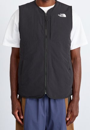 Mężczyzna ubrany w czarną bezrękawnik na zamek błyskawiczny z logo The North Face, na biały t-shirt i niebiesko-brązowe spodnie.