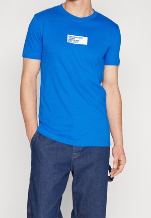 Mężczyzna w jasnoniebieskiej koszulce Calvin Klein z białym logo i ciemnoniebieskich dżinsach, stoi z jedną ręką w kieszeni.