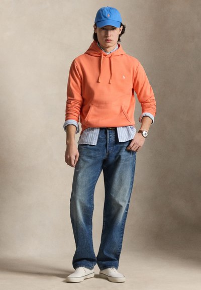 Ung mann med oransje hettegenser, blå stripete skjorte, blå caps, vide blå jeans, hvite joggesko og et armbåndsur, står mot en ensfarget bakgrunn.