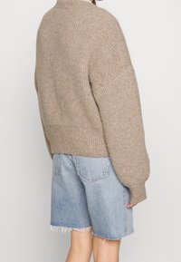 Persona che indossa un maglione beige a costine e pantaloni shorts di jeans azzurri a vita alta con orli sfilacciati, in piedi contro uno sfondo semplice.