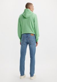 Άνδρας με ανοιχτό πράσινο φούτερ, μπλε τζιν και λευκά αθλητικά παπούτσια, στέκεται με την πλάτη προς το φακό σε λευκό φόντο.