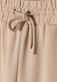 Beige Jogginghose mit einem Kordelzugbund und dekorativem Knoten, glatter Stofftextur und Kunststoffspitzen an den Enden des Kordelzugs.