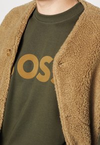Chaqueta de forro polar marrón y esponjosa con cierre de botones sobre un suéter verde oscuro que presenta un gran logo impreso de color mostaza.