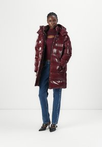 Burgundy puffer coat with a glossy finish, featuring a high collar, zip closure, and oversized pockets, worn over dark jeans and heels.