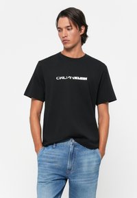 Black cotton t-shirt with short sleeves, round neckline, and white "CALVIN KLEIN" logo on the front; paired with light blue denim jeans.