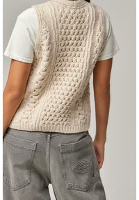 Beige pletená vesta s texturovanými vzory, nošená přes světle šedé tričko. Šedé kalhoty mají velké kapsy a reliéfní logo.