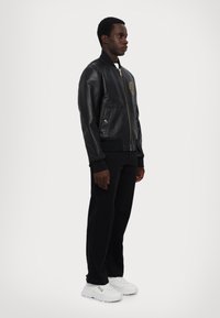 Giubbotto bomber in pelle nera con un emblema dorato, polsini e orlo a coste, abbinato a pantaloni neri e sneakers bianche. Vista laterale.