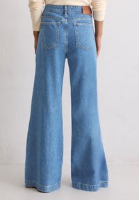 Persoon draagt hoge taille, wijd uitlopende blauwe spijkerbroeken met achterzakken en een tan leren label, gezien van achteren tegen een plain witte muur.