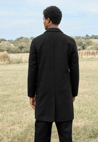 Abrigo de lana negro con un collar estructurado, mangas largas y una abertura en la parte trasera. Destaca por sus líneas limpias y diseño minimalista.
