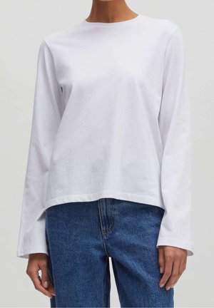 Person trägt ein schlichtes weißes Langarmshirt kombiniert mit blauen Jeans und steht vor einem neutralen Hintergrund.