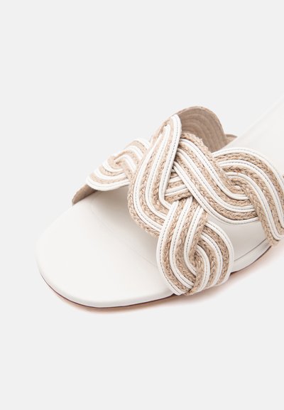 Άσπρο σανδάλι ανοιχτών δαχτύλων με χοντρά, πλεγμένα λουριά από φυσικές ίνες και άσπρα δερμάτινα λουριά που διασταυρώνονται πάνω από το πόδι.