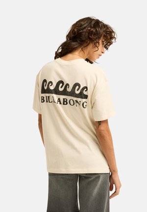 Kobieta z kręconymi włosami w kremowej koszulce Billabong z czarnymi falistymi grafikami na plecach i wyblakłych szarych dżinsach.