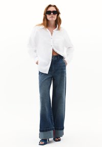 Camicia bianca con bottoni e tasca, abbinata a jeans in denim blu a gamba larga con orlo in contrasto e scarpe col tacco blu.
