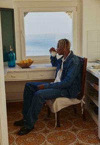 Traper jakna i traperice, bijela košulja, sjedi na drvenoj stolici. Pije iz šalice kraj prozora, s košarom voća i plavom staklenom bocom.