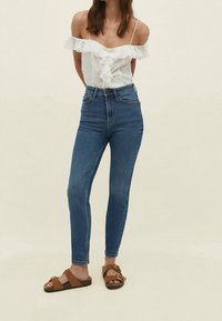 Witte top met blote schouders en ruches, hoge blauwe skinny jeans en bruine sandalen met dubbele gespen. Gladde stof en aansluitend ontwerp.