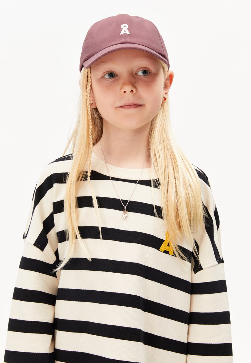 Lila Kappe mit einem weißen Band-Logo, gestreifter oversized Pullover in Schwarz und Creme mit gelbem Buchstaben "A" und silberner Herzanhänger-Halskette.