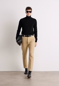 Pulover negru cu gât, pantaloni bej, curea neagră și pantofi negri. Persoană care ține o geantă neagră, poartă ochelari de soare închiși și stă pe un fundal simplu.