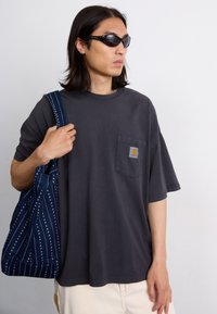 T-shirt oversize gris foncé avec une poche poitrine et un patch logo. La personne porte un sac marine avec des motifs et des rayures blanches.
