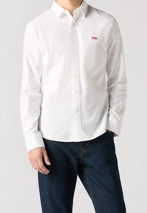 Mand iført en hvid Levi's skjorte med knapper og lange ærmer samt mørkeblå jeans, stående med den ene hånd i lommen.