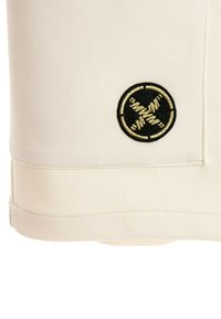 Logotipo redondo bordado en negro y dorado con pesas de barra cruzadas sobre tela crema con costuras visibles cerca del dobladillo.