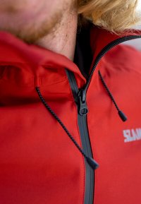 Veste imperméable rouge avec un col haut, fermeture éclair noire, cordons de serrage ajustables et un logo. Caractérisée par un tissu lisse et texturé.