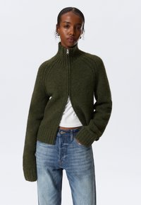 Jersey de cremallera verde oscuro con cuello y puños acanalados. Textura suave, diseño corto, usado sobre una camiseta blanca, combinado con jeans de color azul claro.