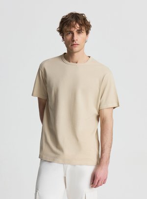 Giovane uomo con capelli ricci che indossa una camicia beige a maniche corte con trama e pantaloni bianchi su uno sfondo chiaro semplice.
