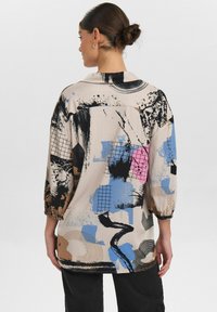 Blouse ample beige avec des motifs abstraits noirs, bleus, roses et marron ; possède un col et des manches trois-quarts.