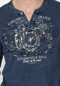 Tamno plavi henley majica s printanim grafikama, dugmadima i kontrastnim šavovima. Tekst uključuje "UNLIMITED BRAND" i "MOTORCYCLE SOUL."