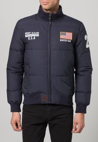 Navy blue puffer jacket with a high collar, zip front, and ribbed cuffs. Features American flag, text patches, and a number "22" on the sleeve.