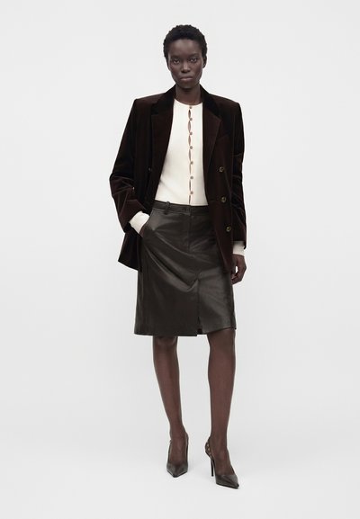 Femme portant un blazer en velours foncé, un pull blanc boutonné, une jupe en cuir noire longueur genou et des chaussures à talons noirs, debout sur un fond blanc.