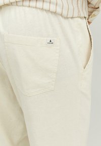 Beige linen pants with visible back pocket featuring a small white Jack & Jones label, worn with a striped beige and white shirt.