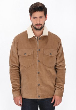 Mand med kort brunt hår og skæg iført en beige fløjlsjakke med hvid sherpa-krave står mod en ensfarvet baggrund.