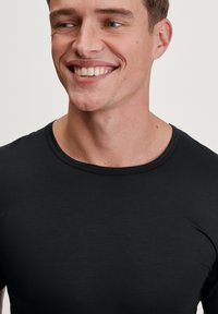 Black short-sleeve crewneck t-shirt displayed on a smiling male model. The fabric appears smooth with a slight sheen.