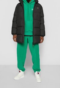 Svart puffjacka med huva, grön hoodie med logotyp, matchande gröna sweatpants och vita sneakers med logotypdetaljer.