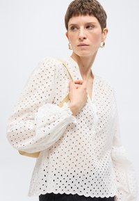 Γυναίκα με κοντά μαλλιά, φοράει λευκό μπλουζάκι με τρυπητό σχέδιο και κυματιστό τελείωμα, κρατάει μπεζ τσάντα ώμου και κοιτάζει στο πλάι με φόντο το λευκό.