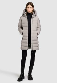 Lys grå pufferjakke med hætte, udstyret med et quiltet design, lynlåslommer og knælang pasform. Båret over en sort turtleneck og tights.
