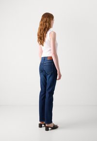 Jeans justos de cor azul escuro com bolsos traseiros, etiqueta de couro na cintura, usados com uma blusa branca sem mangas e mules pretos de salto bloco.