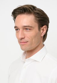 Camicia bianca con colletto strutturato, texture liscia e chiusura con bottoni, indossata da un uomo con capelli castani di lunghezza media e pelle chiara.