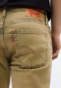 Κοντινό πλάνο ατόμου που φοράει ανοιχτό καφέ τζιν Levi’s, δείχνοντας την πίσω τσέπη, την κόκκινη ετικέτα Levi’s και το δερμάτινο σήμα της μάρκας στη μέση.