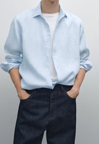 Camicia azzurro chiaro in cotone, con maniche arrotolate e vestibilità rilassata, abbinata a jeans in denim scuro e una maglietta bianca sotto.