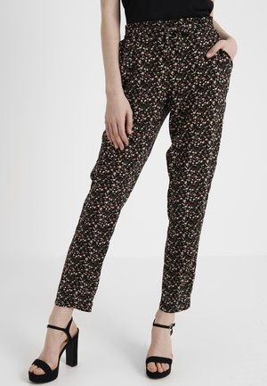 Frau trägt schwarze, floral gemusterte, schmal geschnittene Hose mit elastischem Bund, kombiniert mit schwarzen Sandalen mit Absatz und einem schwarzen Oberteil.