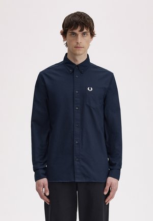 Giovane uomo con capelli corti castani che indossa una camicia a maniche lunghe blu scuro con bottoni e un piccolo logo bianco sulla tasca sul petto.