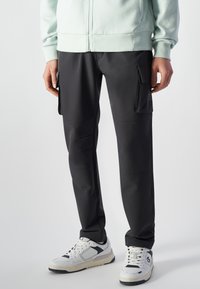 Zwarte cargobroek met meerdere zijzakken, gemaakt van een gladde stof. Gecombineerd met witte en marineblauwe sneakers en een lichtgroene hoodie met rits.