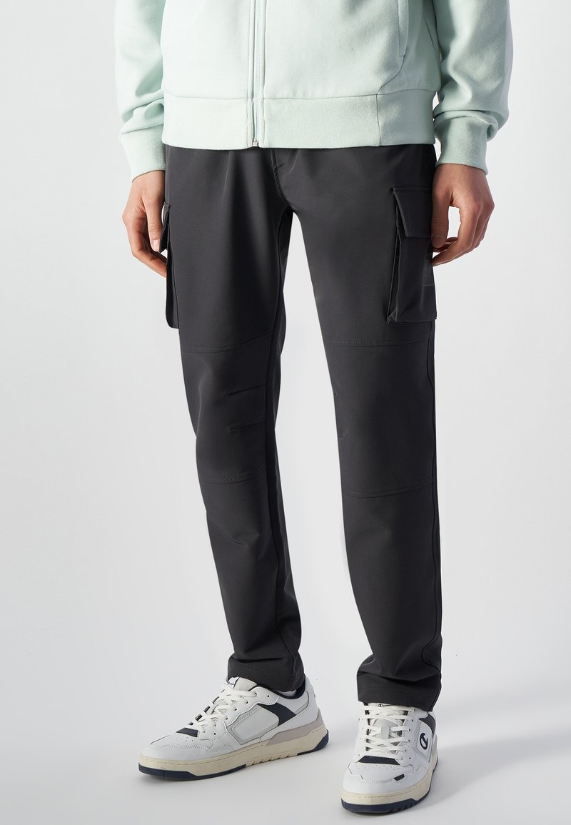 Zwarte cargobroek met meerdere zijzakken, gemaakt van een gladde stof. Gecombineerd met witte en marineblauwe sneakers en een lichtgroene hoodie met rits.