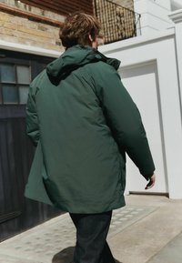 Green padded jacket with a hood, featuring a smooth texture and loose fit, paired with dark denim pants. Texture appears lightweight.