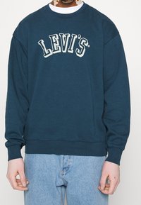Námořnická modrá mikina s kulatým výstřihem, spadlými rameny a bílým logem "LEVI'S" na hrudi. Měkká tkanina s žebrovanými manžetami a lemem.